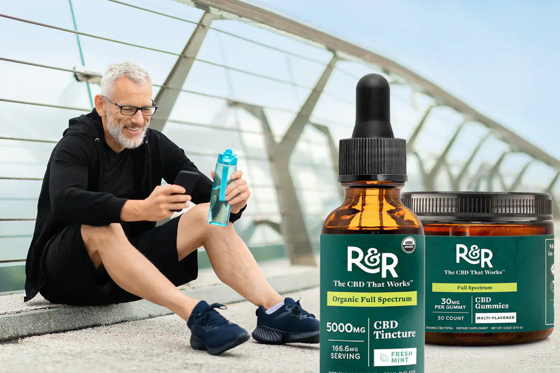 A man is sitting on the roadside, watching something on his mobile, with an R&R CBD Tincture and a CBD Gummies container beside him